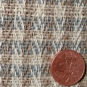 May include: Close-up of woven fabric with a mix of beige, light blue, and brown threads. A copper two pence coin is in the lower right corner, providing a size reference. The fabric has a textured, basket-weave appearance.