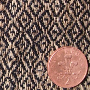May include: Close-up of a textured fabric with a black and beige herringbone pattern. A copper two pence coin is in the foreground, providing a size reference. The fabric appears to be woven, possibly wool or a similar material.