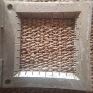May include: Close-up of a woven, natural fiber material, possibly jute or sisal, framed by a metal measuring tool. The texture is coarse and the color is a warm, earthy brown. The measuring tool has etched markings.