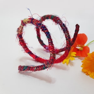 May include: A handmade, multi-colored bracelet with a spiral design. The bracelet is primarily red with accents of blue, green, and gold. The image also includes orange and yellow flowers.