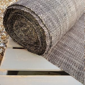 May include: A rolled bolt of fabric with a repeating diamond pattern in black and beige. The fabric is resting on a light gray wooden surface, with the roll's end showing the tightly wound layers.