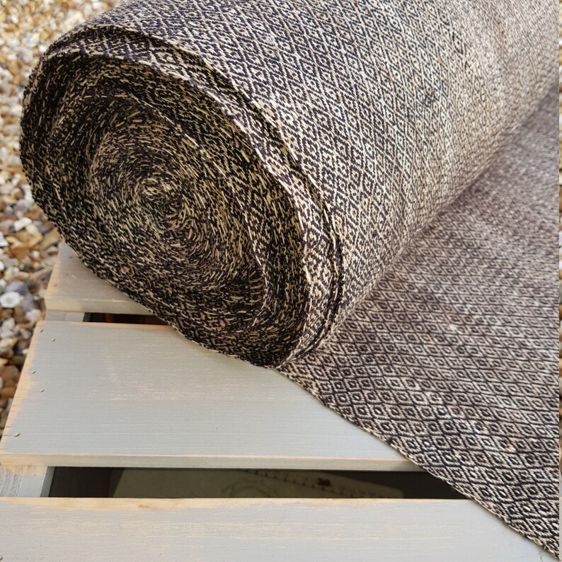 May include: A rolled bolt of fabric with a repeating diamond pattern in black and beige. The fabric is resting on a light gray wooden surface, with the roll's end showing the tightly wound layers.