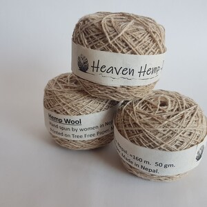 Peut inclure: Trois boules de fil de laine de chanvre de couleur naturelle sont empil&eacute;es. Chaque boule a une bande de papier blanche avec le texte "Heaven Hemp" et "Hemp Wool". Le fil est fil&eacute; &agrave; la main par des femmes au N&eacute;pal.
