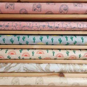 Puede incluir: Una pila de rollos de papel de regalo estampados con varios diseños. Los diseños incluyen cactus, soles y caras abstractas. Los rollos se exhiben en estantes de madera, mostrando los diferentes diseños y colores.