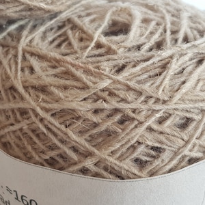 Peut inclure: Gros plan sur une pelote de laine beige, &eacute;troitement enroul&eacute;e avec une texture visible. Le fil semble &ecirc;tre fait de fibres naturelles, adapt&eacute; aux projets de tricot ou de crochet. Une partie d'une &eacute;tiquette est visible.