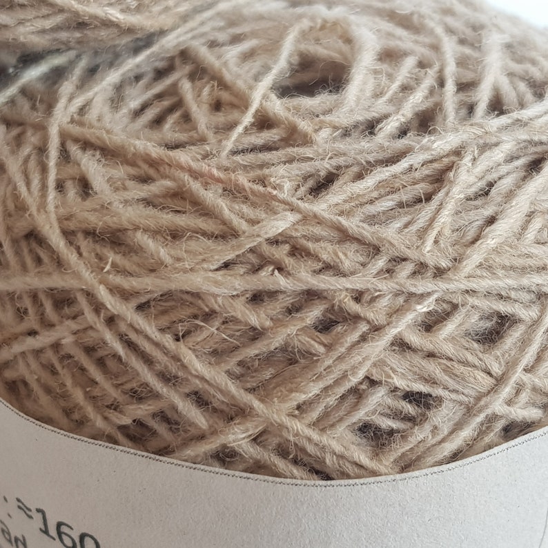 Peut inclure: Gros plan sur une pelote de laine beige, &eacute;troitement enroul&eacute;e avec une texture visible. Le fil semble &ecirc;tre fait de fibres naturelles, adapt&eacute; aux projets de tricot ou de crochet. Une partie d'une &eacute;tiquette est visible.