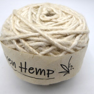 Peut inclure: Une pelote de fil de chanvre tiss&eacute; de couleur naturelle. Le fil est &eacute;troitement enroul&eacute; et a un aspect textur&eacute;. Une &eacute;tiquette en papier enroul&eacute;e autour de la base indique "Woven Hemp" en texte noir, avec une illustration de feuille de cannabis.