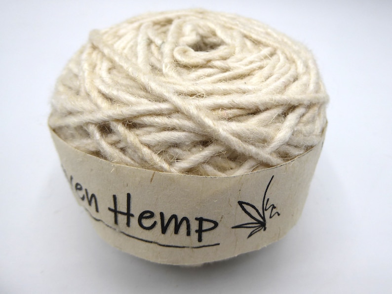 Peut inclure: Une pelote de fil de chanvre tiss&eacute; de couleur naturelle. Le fil est &eacute;troitement enroul&eacute; et a un aspect textur&eacute;. Une &eacute;tiquette en papier enroul&eacute;e autour de la base indique "Woven Hemp" en texte noir, avec une illustration de feuille de cannabis.