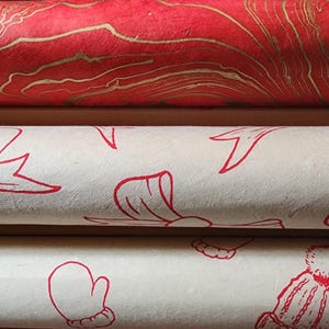 May include: Close-up of three rolls of decorative wrapping paper. The top roll is red with gold marbling. The middle roll is off-white with red bow illustrations. The bottom roll is off-white with red winter hat and mitten illustrations.