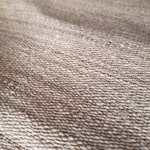 May include: Close-up of a woven fabric with a rough texture. The material is a light brown color, with visible fibers and a slightly uneven surface. The weave pattern is tightly knit, creating a durable appearance.