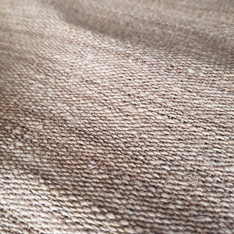 May include: Close-up of a woven fabric with a rough texture. The material is a light brown color, with visible fibers and a slightly uneven surface. The weave pattern is tightly knit, creating a durable appearance.