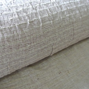 May include: Close-up of a rolled-up, textured fabric in shades of beige and brown. The fabric has a woven appearance with visible fibers and a slightly wrinkled texture. The material appears to be natural, possibly linen or cotton.