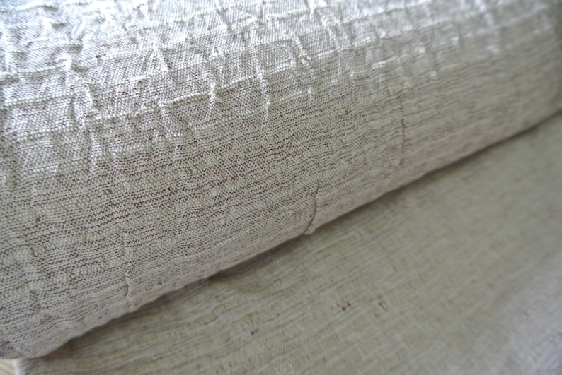 May include: Close-up of a rolled-up, textured fabric in shades of beige and brown. The fabric has a woven appearance with visible fibers and a slightly wrinkled texture. The material appears to be natural, possibly linen or cotton.