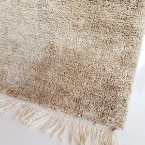 Op de afbeelding: Close-up van een beige tapijt met een harige textuur. Het tapijt heeft een cr&egrave;mekleurige franje langs &eacute;&eacute;n rand. De vezels van het tapijt zijn een mix van lichte en medium beige tinten, waardoor een textuur ontstaat. Het tapijt is waarschijnlijk gemaakt van een zacht materiaal.