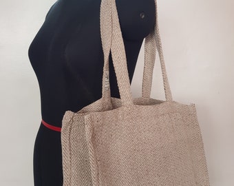 May include: A beige tote bag with a herringbone pattern. The bag has two long handles and is displayed on a black mannequin. The bag is made of a woven fabric and has a tag attached to one of the handles.
