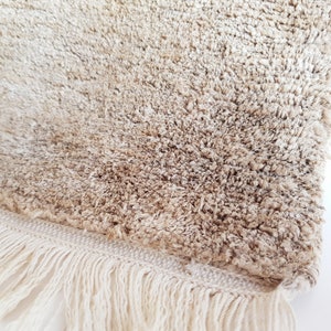 Op de afbeelding: Close-up van een beige, getextureerd tapijt met een harige pool. Het tapijt heeft een cr&egrave;mekleurige franje langs &eacute;&eacute;n rand. De vezels zijn een mix van lichte en medium bruine tinten, wat een zachte uitstraling cre&euml;ert.