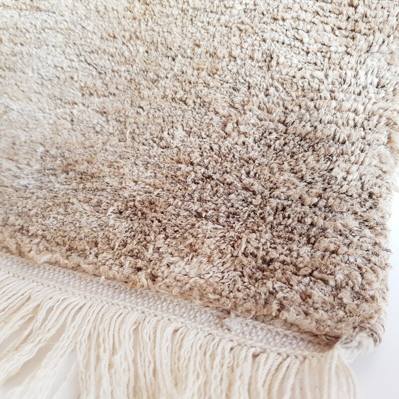 Op de afbeelding: Close-up van een beige, getextureerd tapijt met een harige pool. Het tapijt heeft een cr&egrave;mekleurige franje langs &eacute;&eacute;n rand. De vezels zijn een mix van lichte en medium bruine tinten, wat een zachte uitstraling cre&euml;ert.