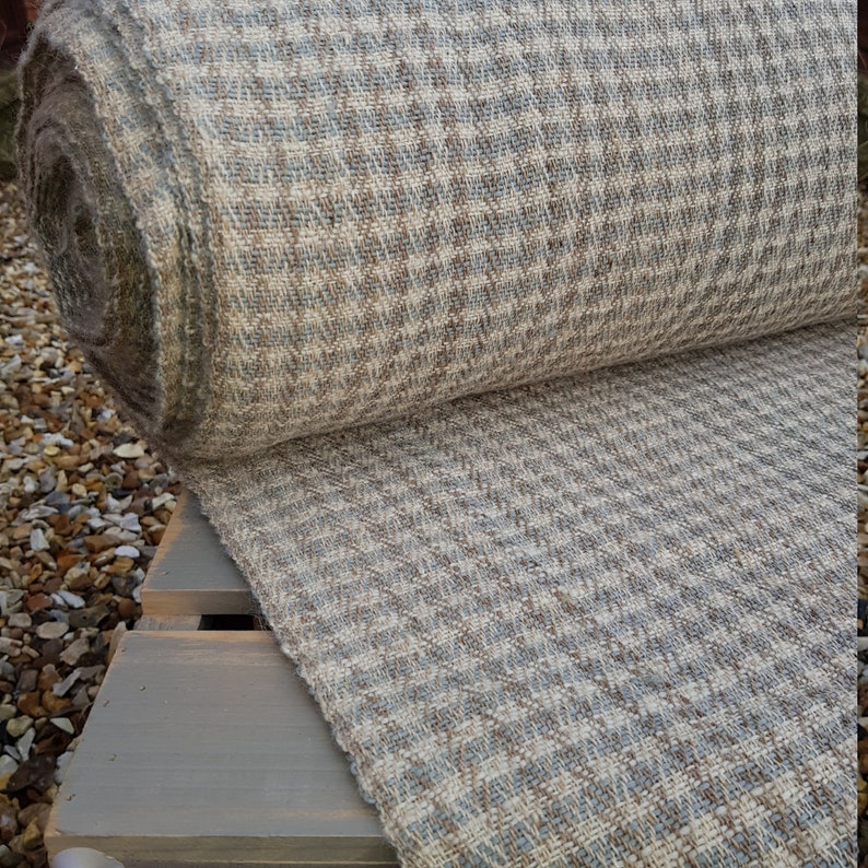 May include: A rolled-up bolt of fabric with a houndstooth pattern in shades of beige, brown, and blue. The fabric is textured and appears to be a woven material, possibly wool or a wool blend. The fabric is resting on a gray wooden surface.