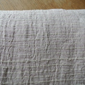 May include: Close-up of a folded, textured fabric in a muted gray-beige color. The material has a woven appearance with horizontal and vertical lines creating a grid-like pattern. The fabric appears soft and slightly wrinkled.