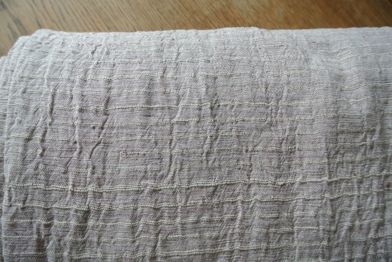 May include: Close-up of a folded, textured fabric in a muted gray-beige color. The material has a woven appearance with horizontal and vertical lines creating a grid-like pattern. The fabric appears soft and slightly wrinkled.