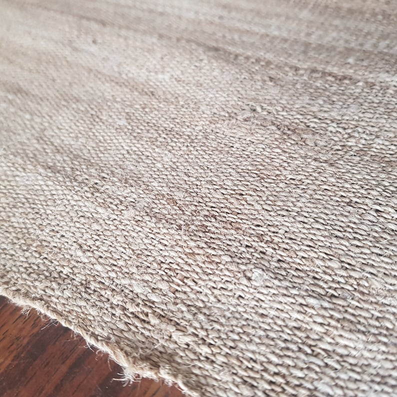 May include: Close-up of a woven jute rug with a natural, light brown color. The rug has a textured surface with visible fibers and a slightly frayed edge. The weave pattern is tight and consistent, creating a durable and rustic aesthetic.