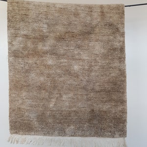 Op de afbeelding: Een rechthoekig, beige tapijt met een getextureerd oppervlak en franjes langs de onderrand. Het tapijt hangt tegen een witte achtergrond en toont zijn neutrale kleur en geweven ontwerp. De afmetingen van het tapijt zijn niet zichtbaar.