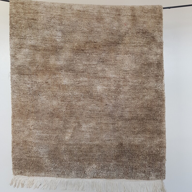 Op de afbeelding: Een rechthoekig, beige tapijt met een getextureerd oppervlak en franjes langs de onderrand. Het tapijt hangt tegen een witte achtergrond en toont zijn neutrale kleur en geweven ontwerp. De afmetingen van het tapijt zijn niet zichtbaar.