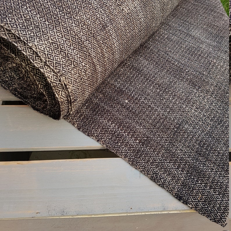 May include: A roll of fabric with a repeating diamond pattern in black and beige. The fabric is partially unrolled, revealing the pattern and texture. The fabric appears to be a medium weight, suitable for various sewing projects.