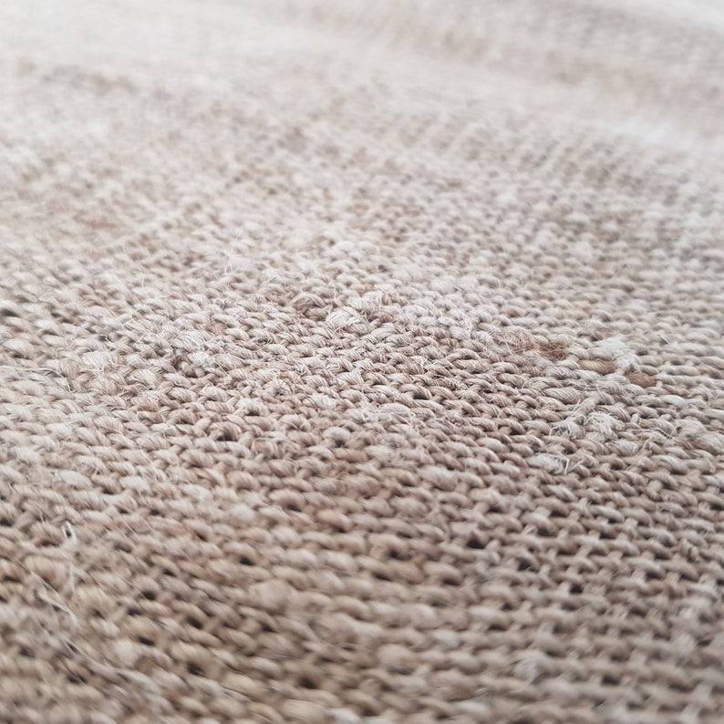 May include: Close-up of a woven textile with a natural, beige color. The texture is rough and appears to be made of thick, intertwined fibers, creating a pattern of small, irregular knots. The focus is on the detailed surface of the fabric.