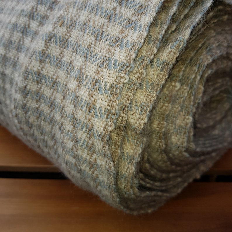 May include: Rolled-up woven fabric in shades of beige, blue, and brown. The textile has a textured appearance with a subtle pattern. The fabric is tightly woven, suggesting durability and a natural fiber composition. The roll displays the fabric's layered construction.