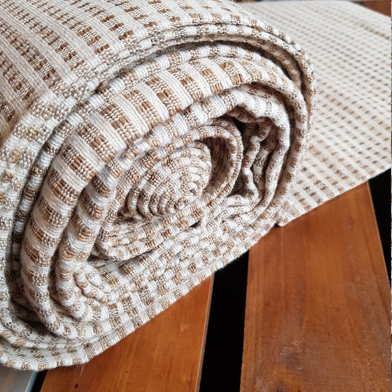 May include: Rolled-up beige and brown woven fabric with a textured, grid-like pattern. The fabric is tightly rolled, revealing the interior layers and the detailed weave. The material appears soft and has a natural, earthy tone.