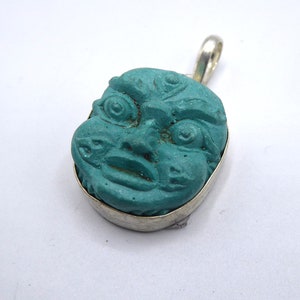 May include: A turquoise pendant with a carved face, set in a silver-coloured metal frame. The face has detailed features, including large eyes and a smiling mouth. The pendant has a loop for a chain. The pendant is a unique piece of jewellery.