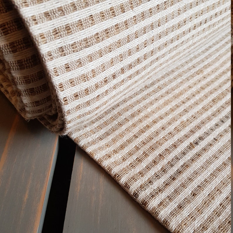 May include: Close-up of a folded, textured fabric with a striped pattern. The fabric features alternating bands of beige and off-white, creating a woven appearance. The material appears to be natural, possibly linen or cotton.