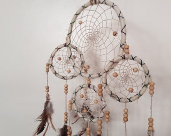 May include: A handmade dreamcatcher with four circular woven frames of varying sizes. The frames are wrapped in cream-colored yarn and adorned with wooden beads, feathers, and a central web design. The dreamcatcher hangs from a natural fiber cord.