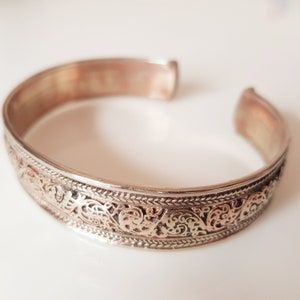 May include: Silver-toned cuff bracelet with intricate, openwork filigree design. The bracelet features a detailed pattern of swirling floral and scroll motifs, creating a delicate and ornate appearance. The open design allows for easy wear and a comfortable fit.