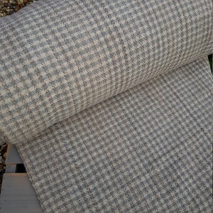 May include: A roll of houndstooth fabric in shades of gray, beige, and blue. The fabric has a woven texture and a classic checkered pattern. The roll is partially unrolled, revealing the fabric's surface and the pattern's detail.