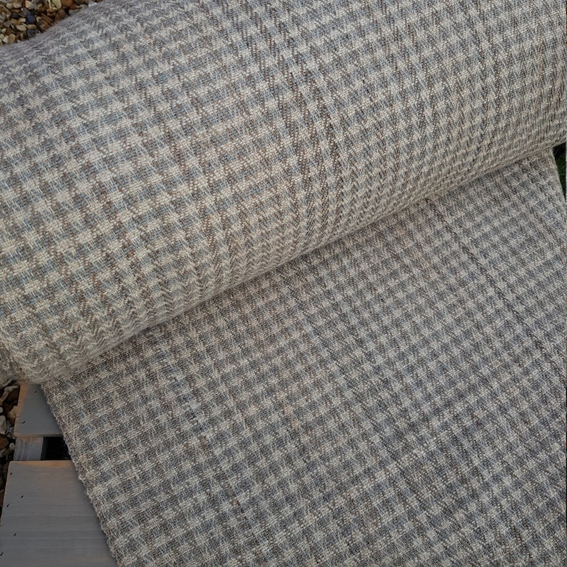 May include: A roll of houndstooth fabric in shades of gray, beige, and blue. The fabric has a woven texture and a classic checkered pattern. The roll is partially unrolled, revealing the fabric's surface and the pattern's detail.