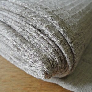 May include: Close-up of a folded stack of light gray linen fabric. The fabric has a textured, ribbed appearance with subtle variations in color. The folds create a layered effect, highlighting the material's texture and drape.