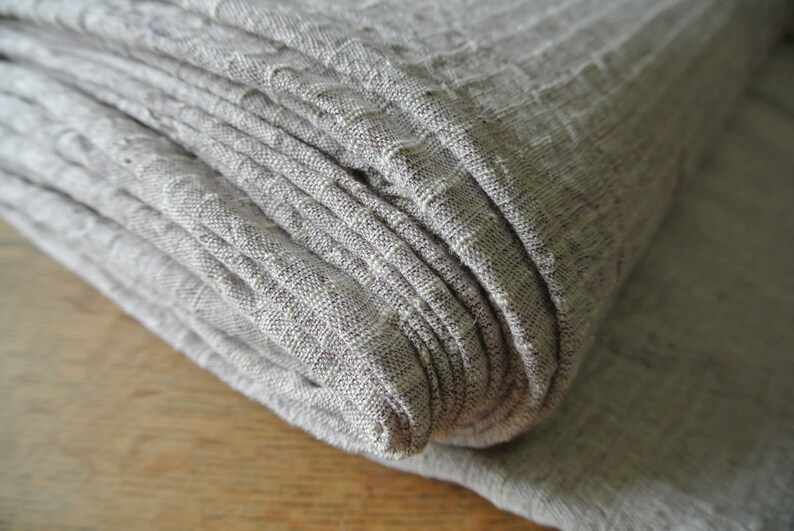 May include: Close-up of a folded stack of light gray linen fabric. The fabric has a textured, ribbed appearance with subtle variations in color. The folds create a layered effect, highlighting the material's texture and drape.