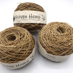 May include: Three balls of natural-coloured hemp yarn. Each ball is wrapped with a paper label that reads "Heaven Hemp" and "Hemp hand spun by women". The yarn has a textured, fibrous appearance, suitable for crafting and fibre arts.