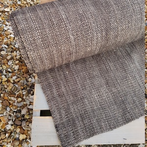 May include: A rolled-up, brown and beige patterned fabric with a diamond design. The fabric is resting on a white wooden pallet, with small rocks in the background. The pattern is repeated across the entire surface.