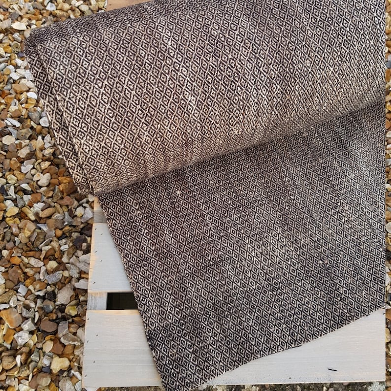May include: A rolled-up, brown and beige patterned fabric with a diamond design. The fabric is resting on a white wooden pallet, with small rocks in the background. The pattern is repeated across the entire surface.
