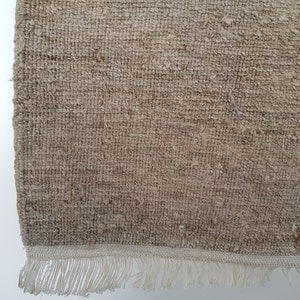 Op de afbeelding: Close-up van een beige geweven vloerkleed met een getextureerd oppervlak. Het vloerkleed heeft een franjeborder in een lichtere tint beige. De weving is zichtbaar en toont de natuurlijke vezels.
