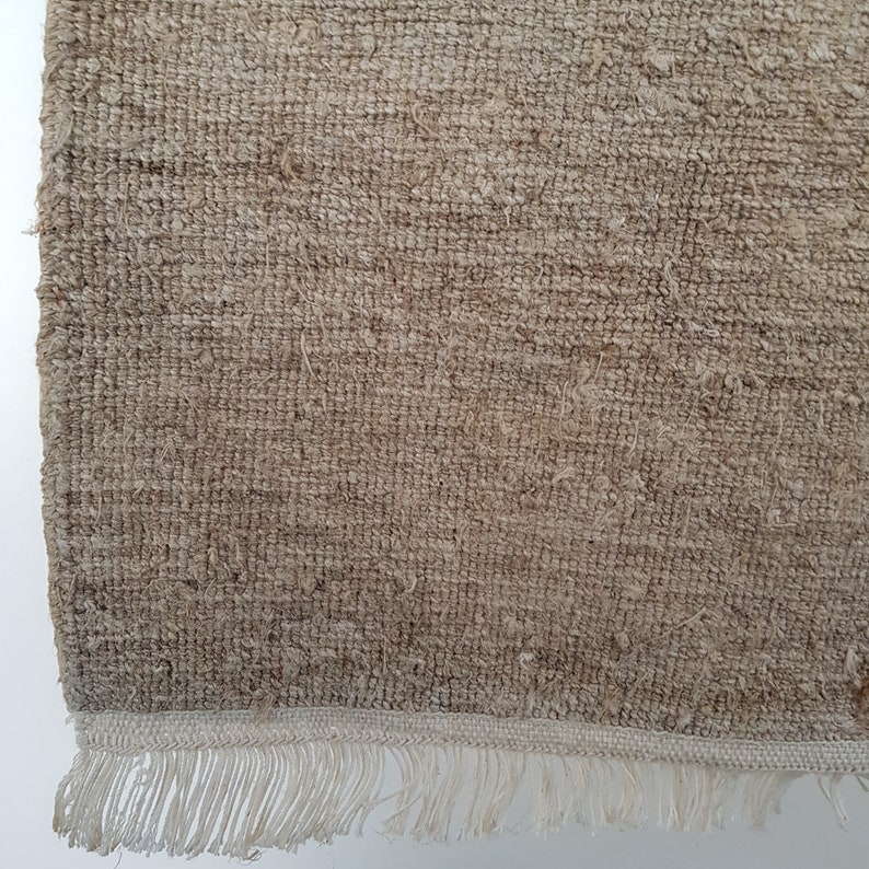 Op de afbeelding: Close-up van een beige geweven vloerkleed met een getextureerd oppervlak. Het vloerkleed heeft een franjeborder in een lichtere tint beige. De weving is zichtbaar en toont de natuurlijke vezels.