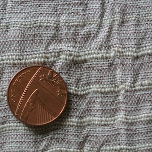 May include: A close-up of a British one penny coin, made of copper, resting on a textured, woven fabric. The coin features the text "ONE PENNY" and a depiction of a ship. The fabric has a beige and maroon color scheme.