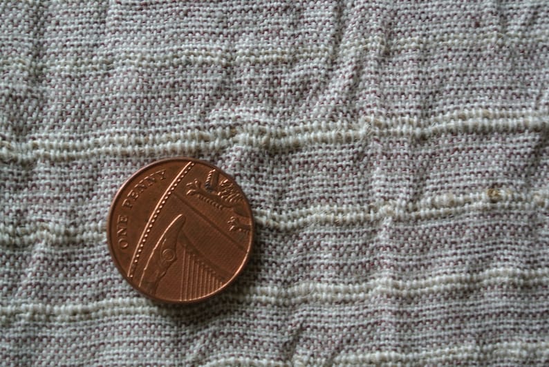 May include: A close-up of a British one penny coin, made of copper, resting on a textured, woven fabric. The coin features the text "ONE PENNY" and a depiction of a ship. The fabric has a beige and maroon color scheme.