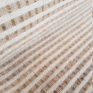 May include: Close-up of a woven fabric with a striped pattern. The fabric features alternating bands of beige and off-white, creating a textured surface. The weave appears to be made of natural fibers, giving it a rustic look.