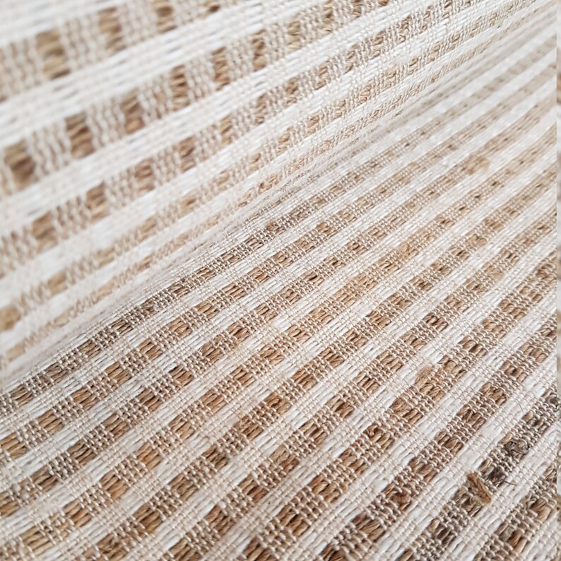 May include: Close-up of a woven fabric with a striped pattern. The fabric features alternating bands of beige and off-white, creating a textured surface. The weave appears to be made of natural fibers, giving it a rustic look.