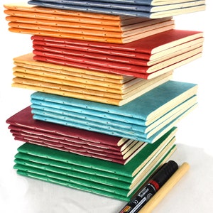 Puede incluir: Una pila de cuadernos hechos a mano en varios colores: azul, naranja, rojo, amarillo, turquesa, burdeos y verde. Cada cuaderno tiene una encuadernación cosida y páginas color crema. Un rotulador negro con mango de madera descansa al lado.