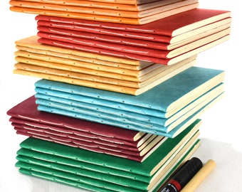 May include: A stack of colorful handmade notebooks in shades of blue, orange, red, yellow, teal, burgundy, and green. Each notebook has a stitched spine and cream-colored pages. A black marker with a wooden handle rests beside the stack.